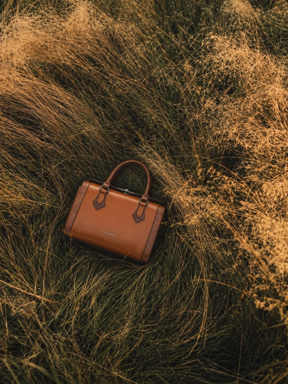 Brown SU QUEENA 2813 bag resting among dry golden meadow grasses, photographed from above.