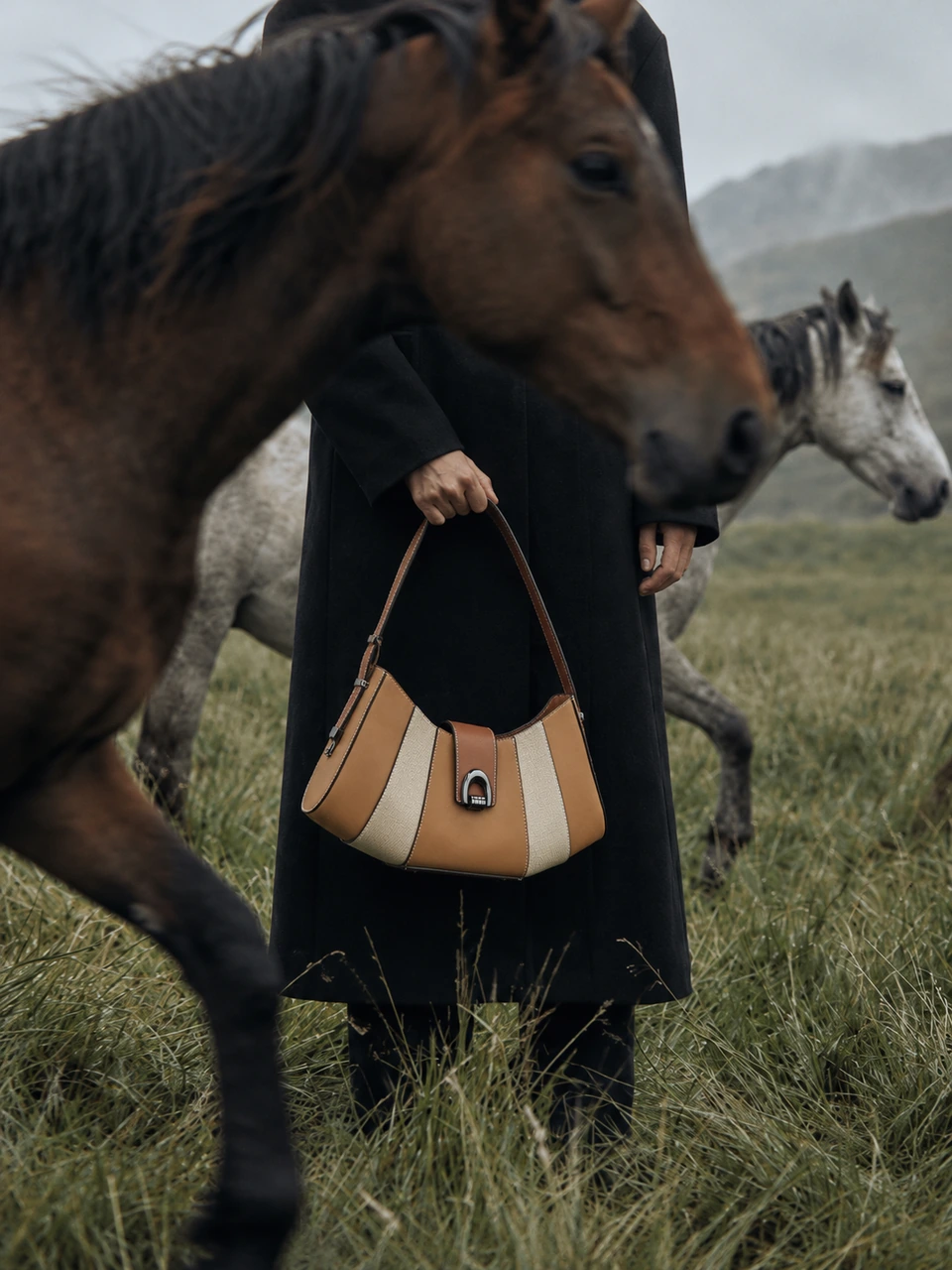 SU QUEENA brown and ivory shoulder bag carried beside horses in a misty meadow.