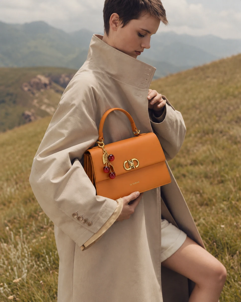 Model in a beige trench holding the SU QUEENA 3244 orange bag with cherry charm in a misty hillside.